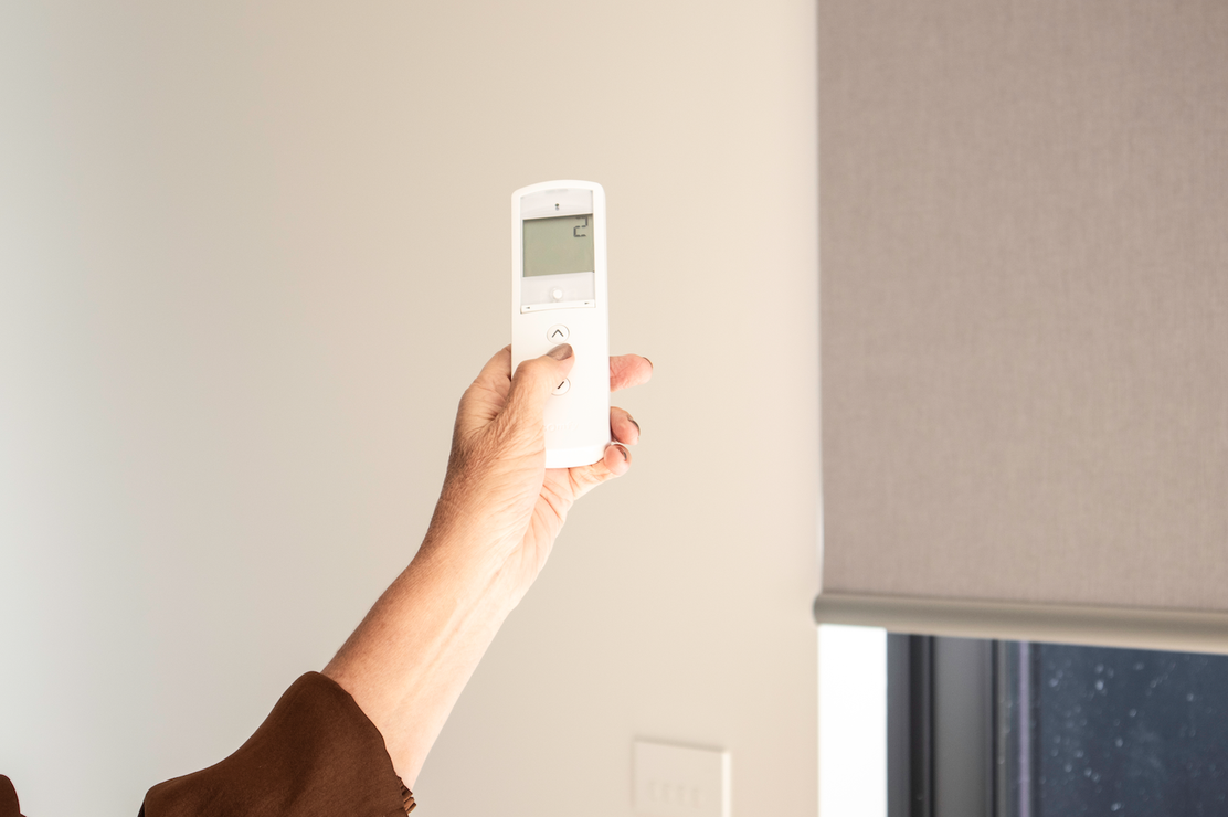 A person holds a remote control towards a window with a closed roller blind, adjusting its position.