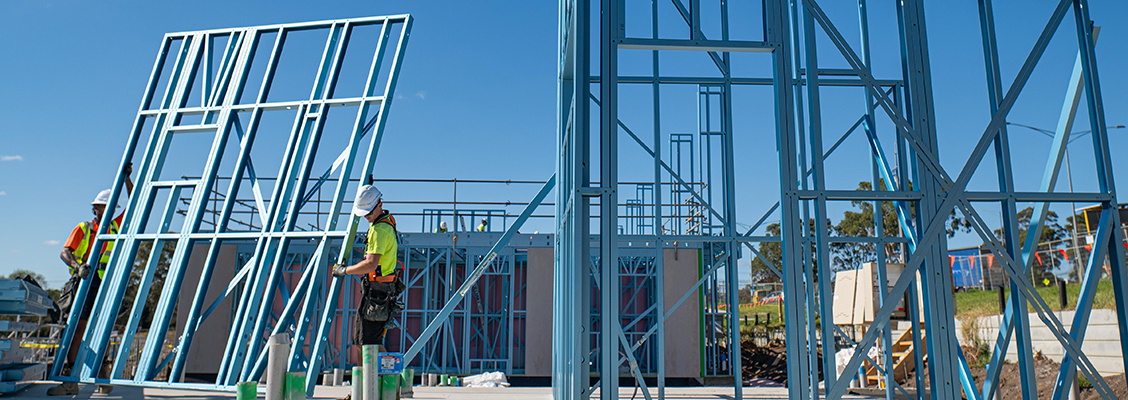 Building site with tradies carrying blue TRUECORE steel frames under a clear blue sky.