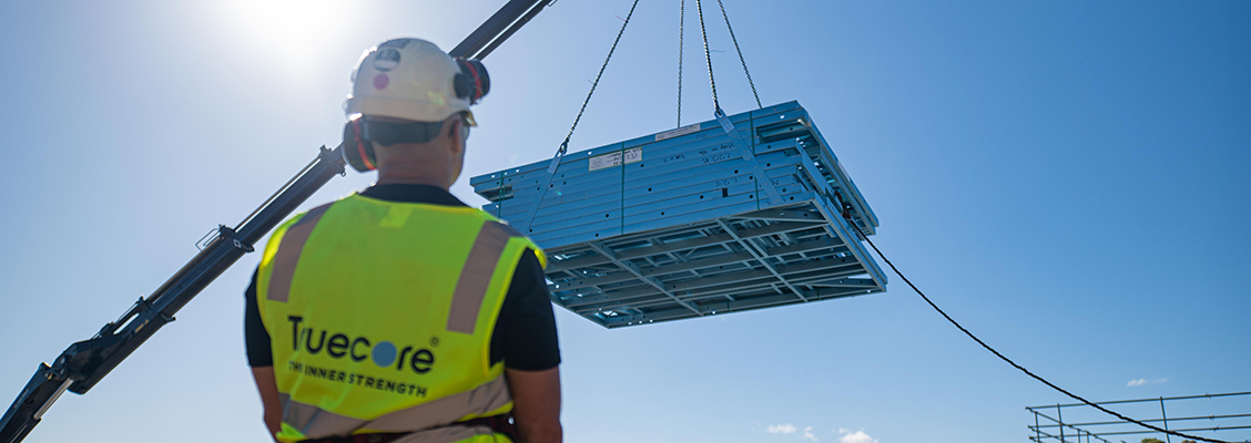 Builder in a yellow vest with TRUECORE steel on it watches as blue TRUECORE steel wall frames are lifted by a crane against a clear blue sky.
