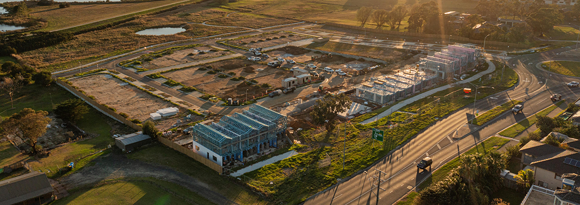 Aerial view of a Creation Homes housing development under construction, with partially built steel framed homes, roads, and surrounding greenery at sunset.