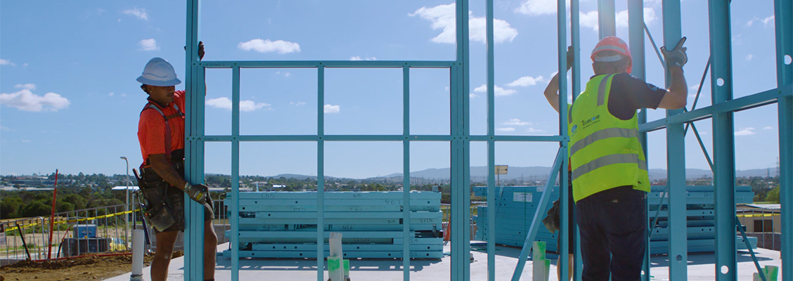 Two construction workers in safety gear carry a blue truecore steel wall frame on a building site under a clear blue sky.