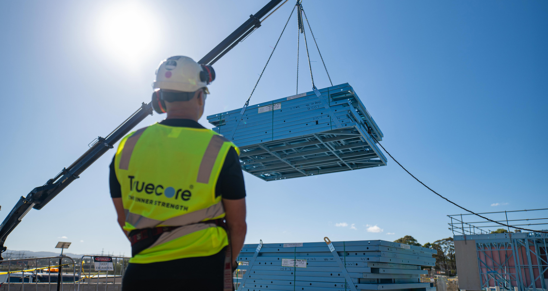 Builder watches a crane lift blue TRUECORE steel wall frames at a construction site under a clear blue sky.