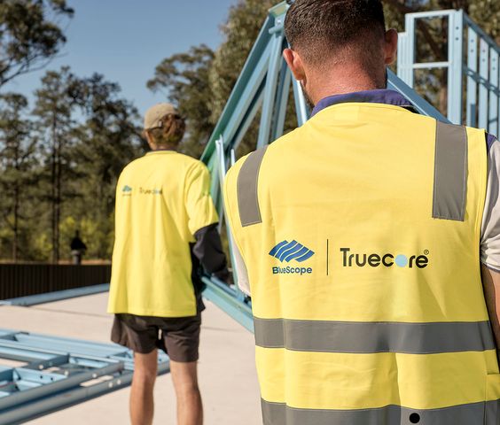 Two workers wearing yellow high-visibility vests with BlueScope and Truecore logos are seen handling a blue steel frame structure outdoors
