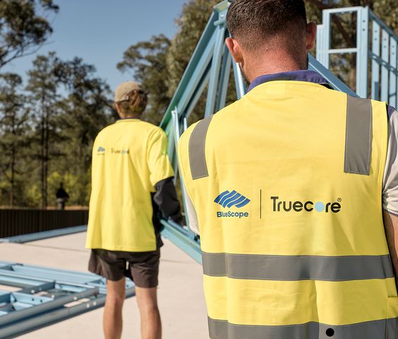 Two workers wearing yellow high-visibility vests with BlueScope and Truecore logos are seen handling a blue steel frame structure outdoors