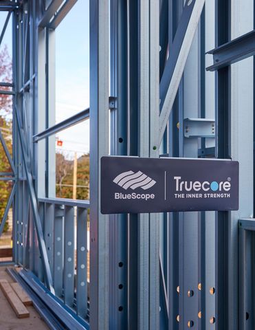 two construction workers wearing yellow safety vests and navy blue hats with the Truecore logo, working together on a metal frame structure.