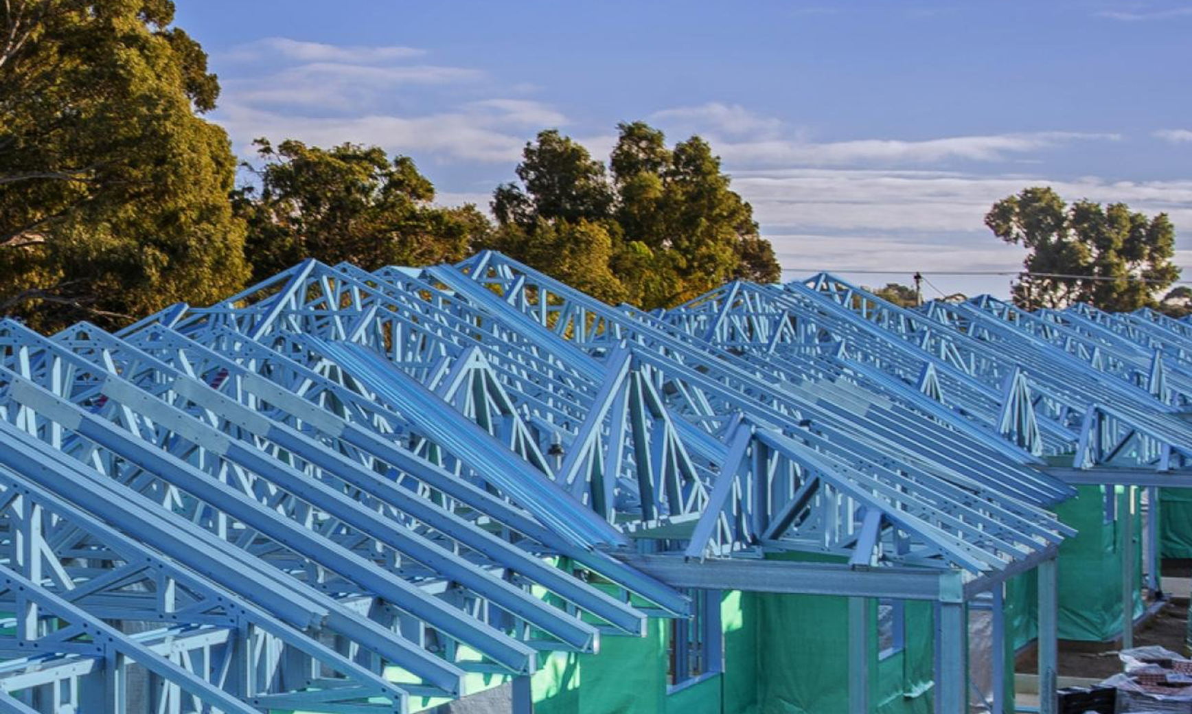 Construction site with several houses in progress, featuring truecore steel blue roof frames and surrounded by trees under a partly cloudy sky.