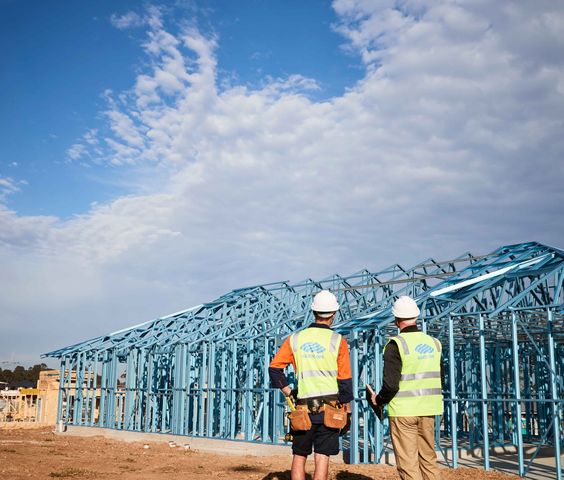 Two construction workers in safety vests and helmets examining the steel framework of a building under construction, with a partly cloudy sky in the background.