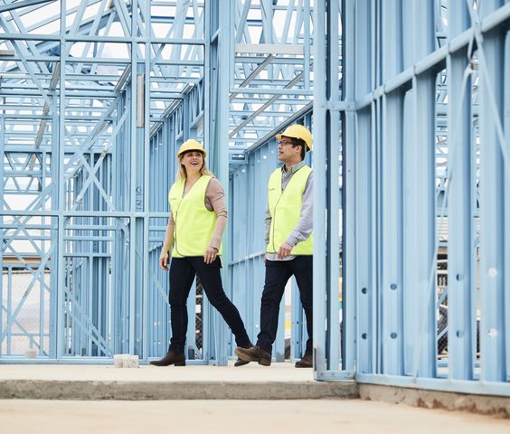 Two people wearing yellow hard hats and reflective vests walk through a construction site framed with blue steel structures