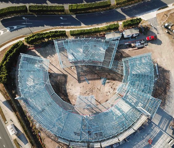 Top-down aerial view of a circular steel-framed building under construction, showing the structure's outline and surrounding area.