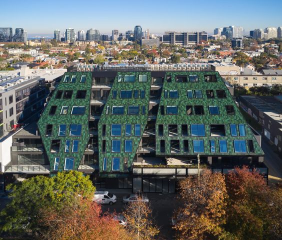 Aerial view of a modern green-roofed building surrounded by urban landscape and trees, with city skyline in the background.