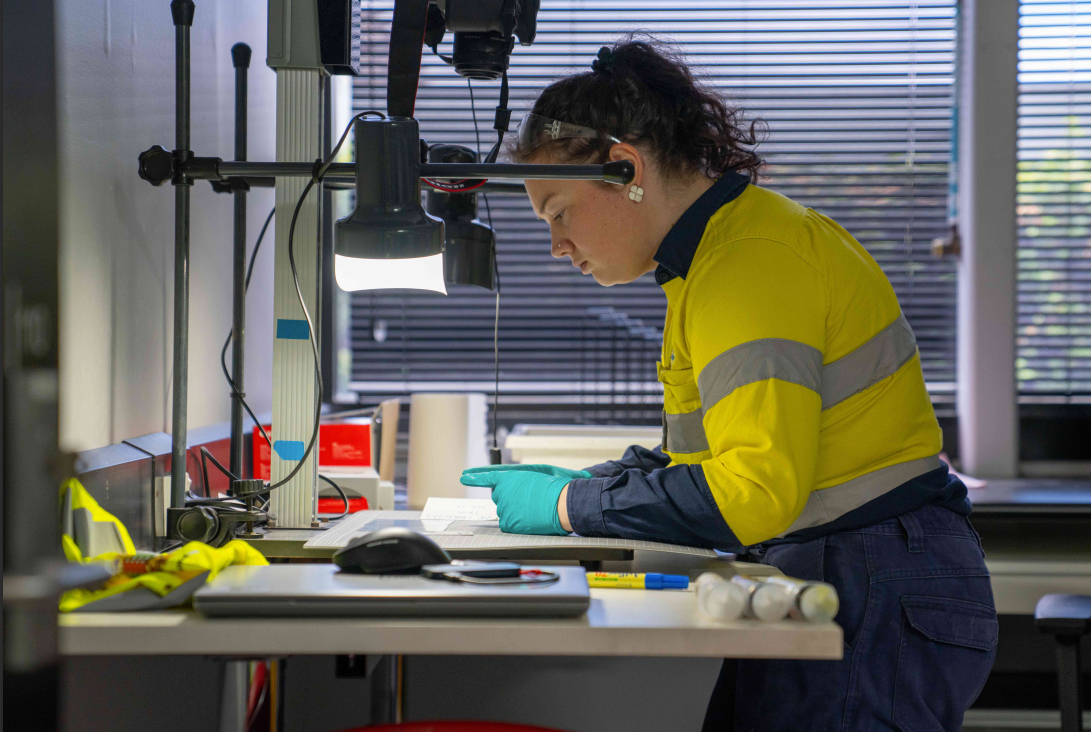 person wearing a high-visibility yellow safety shirt with reflective stripes, working intently at a desk under a bright task light.