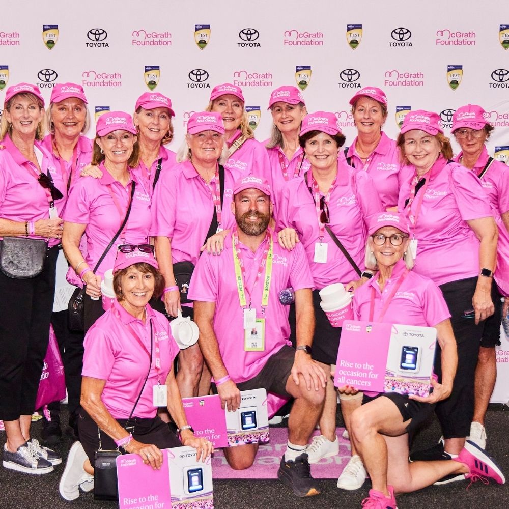 Group of McGrath Foundation volunteers in pink polos and caps posing at a Pink Test event, several kneeling with signs reading "Rise to the occasion for all cancers."