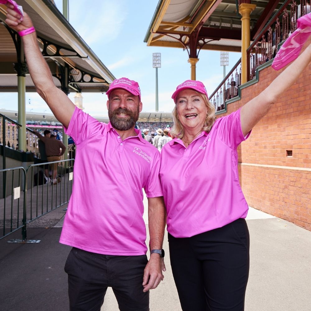 Man and woman in pink McGrath Foundation shirts cheering with raised arms and pink flags at Sydney Cricket Ground.