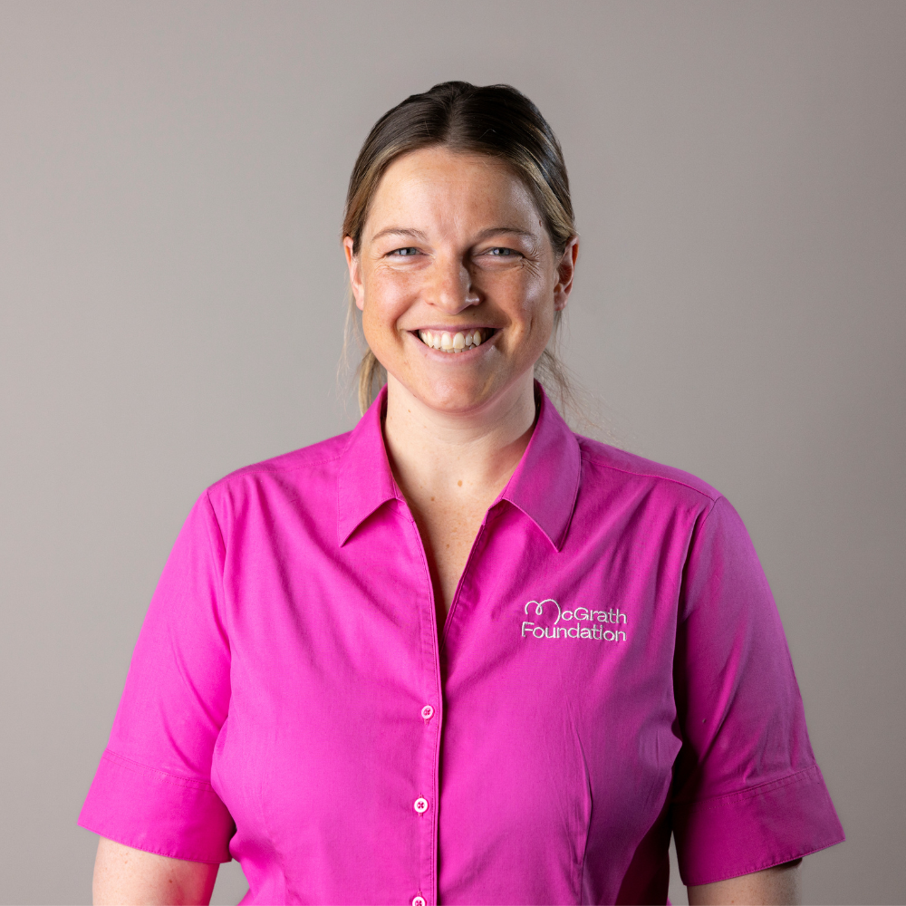 Studio headshot of Elle Conrick in a bright pink button-up shirt with the McGrath Foundation logo, facing forward and smiling, on a plain light grey background with even lighting.