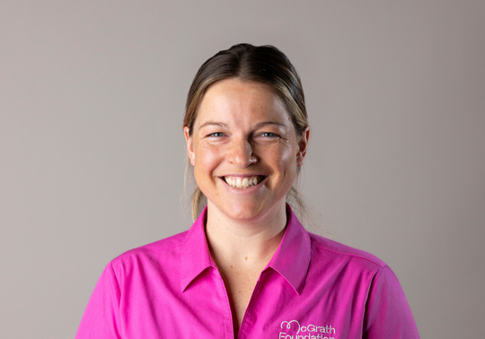 Studio headshot of Elle Conrick in a bright pink button-up shirt with the McGrath Foundation logo, facing forward and smiling, on a plain light grey background with even lighting.