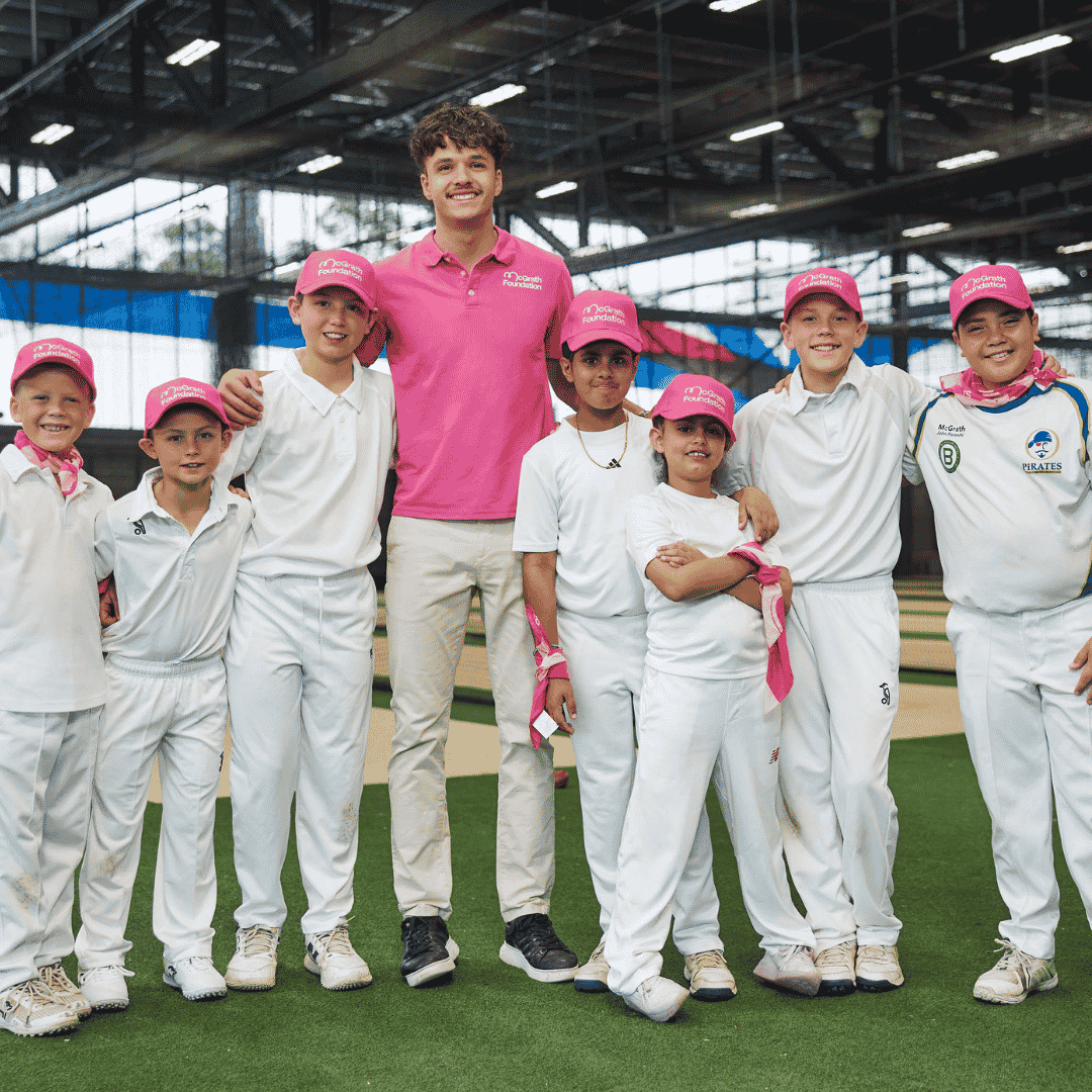 Group of young cricket players wearing pink McGrath Foundation caps stand with Sam Konstas in a pink polo shirt at an indoor cricket facility.