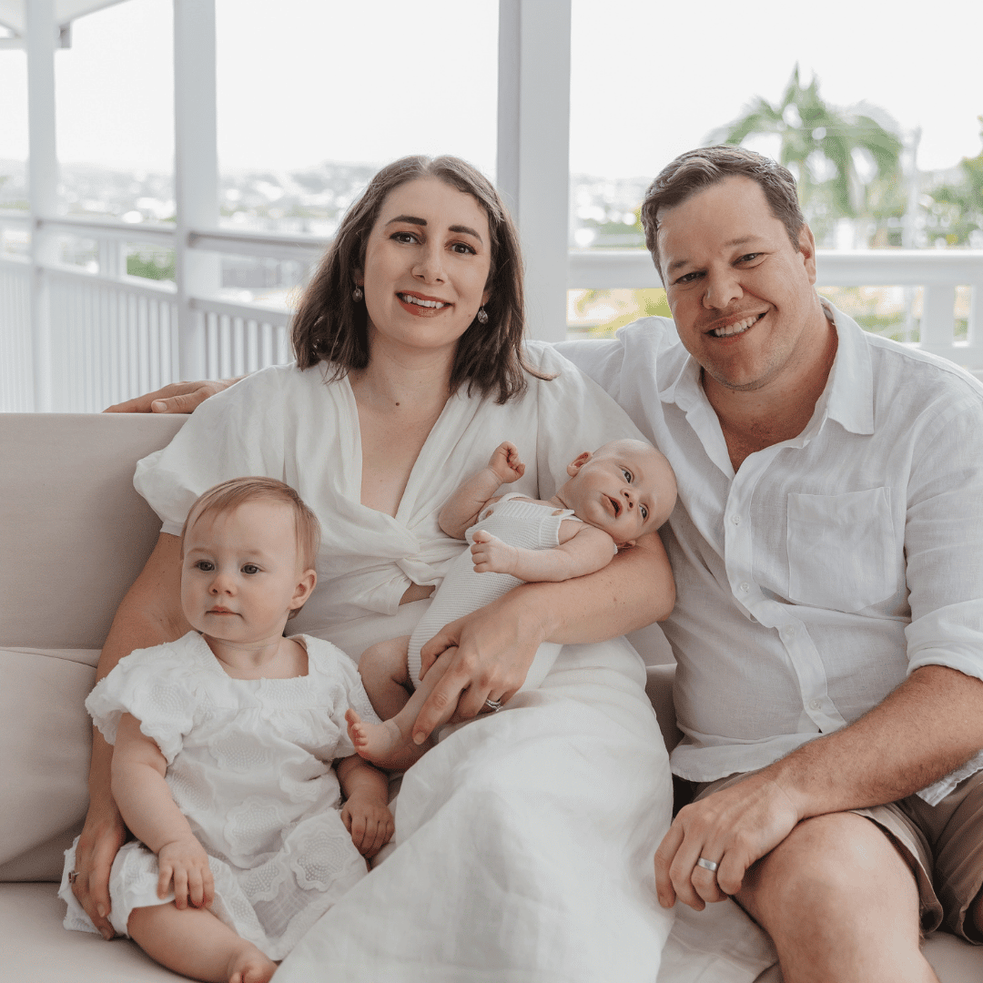 Family of four sitting together on a couch, with two adults holding a baby while a young child sits beside them.
