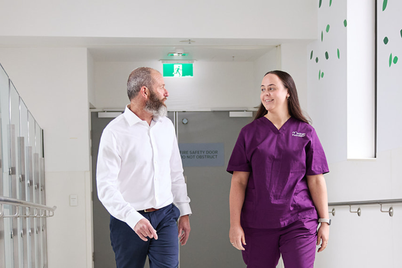 Male patient and his McGrath Cancer Care Nurse walking together down the hallway of a health care facility talking to each other