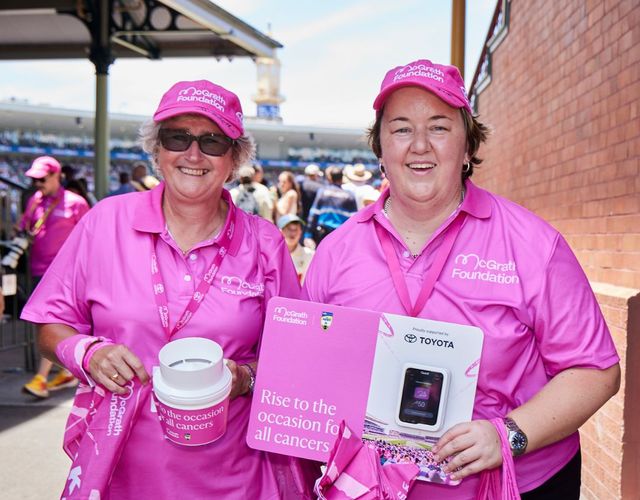 Two McGrath Foundation volunteers in pink shirts hold fundraising materials, donation collection buckets and pink merchandise at a community fundraising event