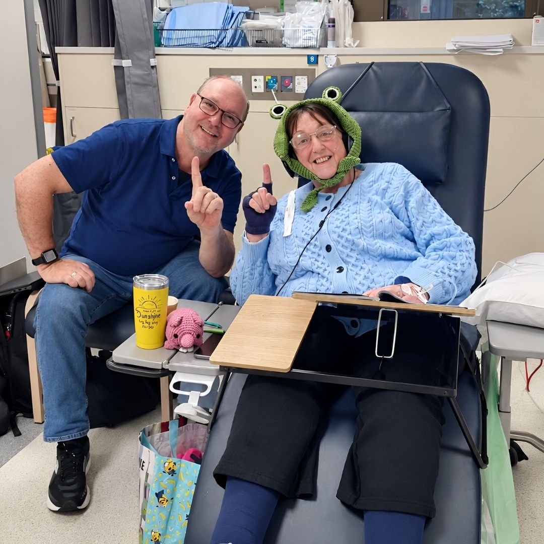 Two people seated together in a hospital treatment room, one wearing a green frog hat