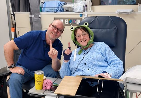 Two people seated together in a hospital treatment room, one wearing a green frog hat