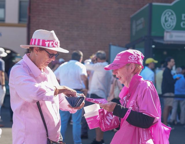 Community member makes a donation to a McGrath Foundation volunteer carrying a donation bucket, event merchandise and fundraising materials.