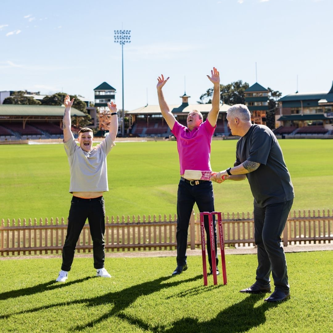 Three people taking part in a cricket-themed photo on a cricket field with pink McGrath Foundation stumps.