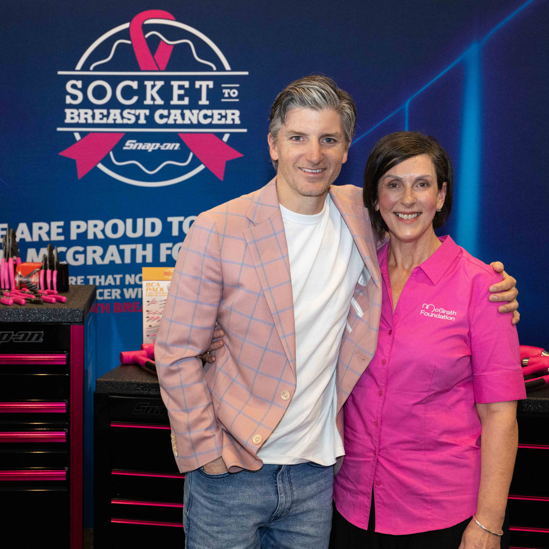 Two people pose together at a Socket Breast Cancer display, with tools and signage in the background and one wearing a pink McGrath Foundation shirt.