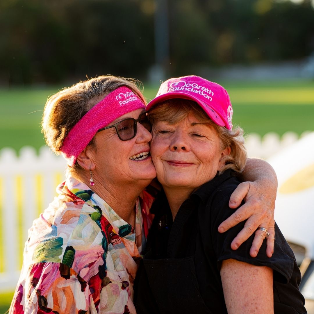Two participants wearing pink McGrath Foundation accessories share a hug at a community fundraising event outdoors.