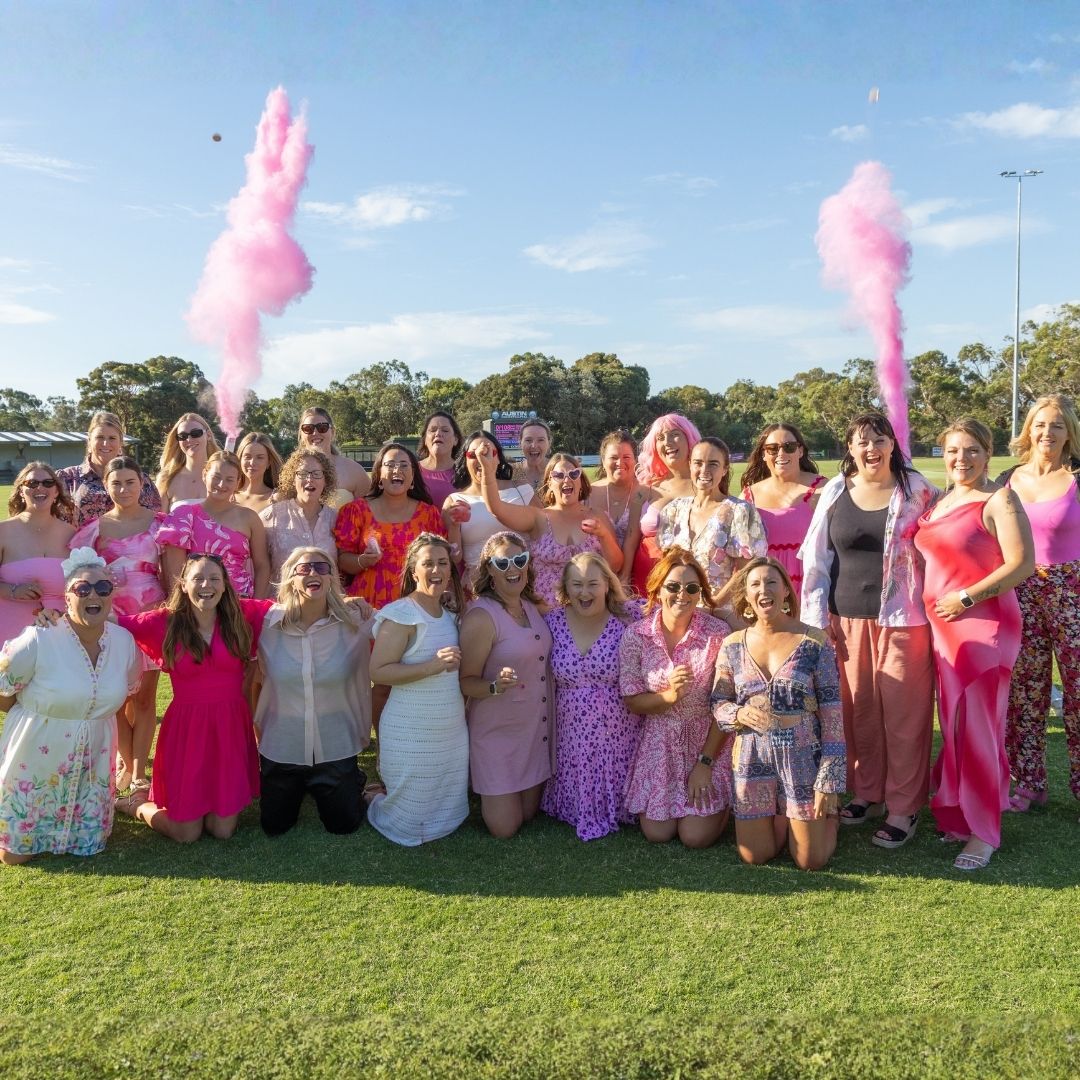Group of people dressed in pink gather on a grassy field during a community fundraising event, with pink smoke flares in the background to show support for all cancers.