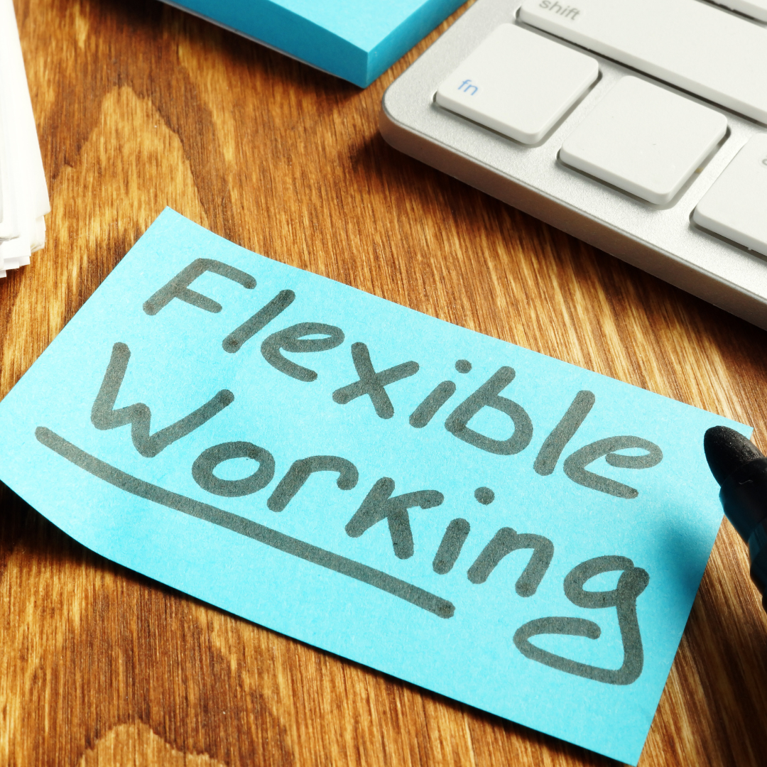 Blue sticky note reading “Flexible Working” on a wooden desk next to a keyboard and marker.