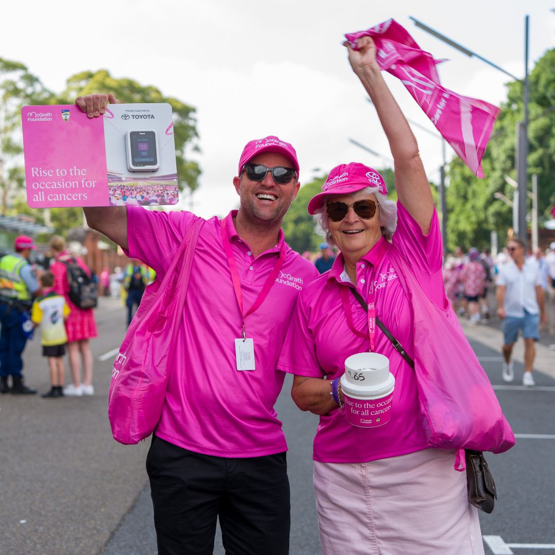 Two volunteers in pink McGrath Foundation shirts stand on an event route holding fundraising materials and waving a pink flag during a cancer fundraising event.