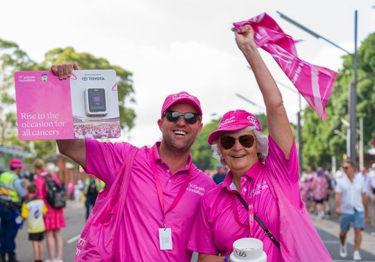Two volunteers in pink McGrath Foundation shirts stand on an event route holding fundraising materials and waving a pink flag during a cancer fundraising event.