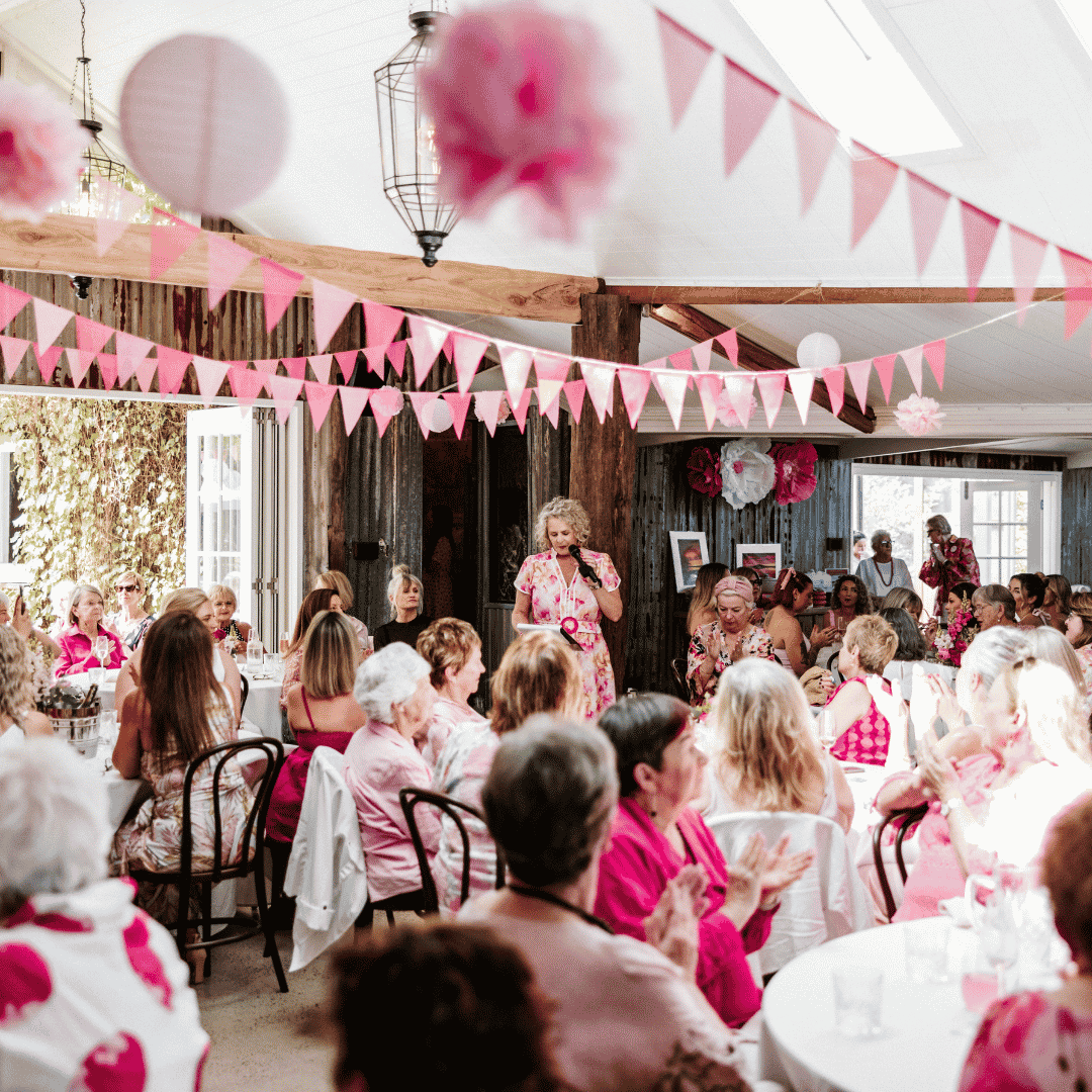 People gathered at a community fundraising event decorated with pink bunting and decorations, with a speaker addressing attendees.