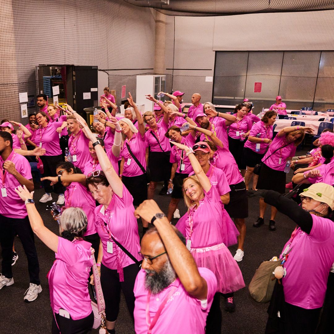 Participants in pink event shirts take part in a group warm-up session indoors before a cancer fundraising event