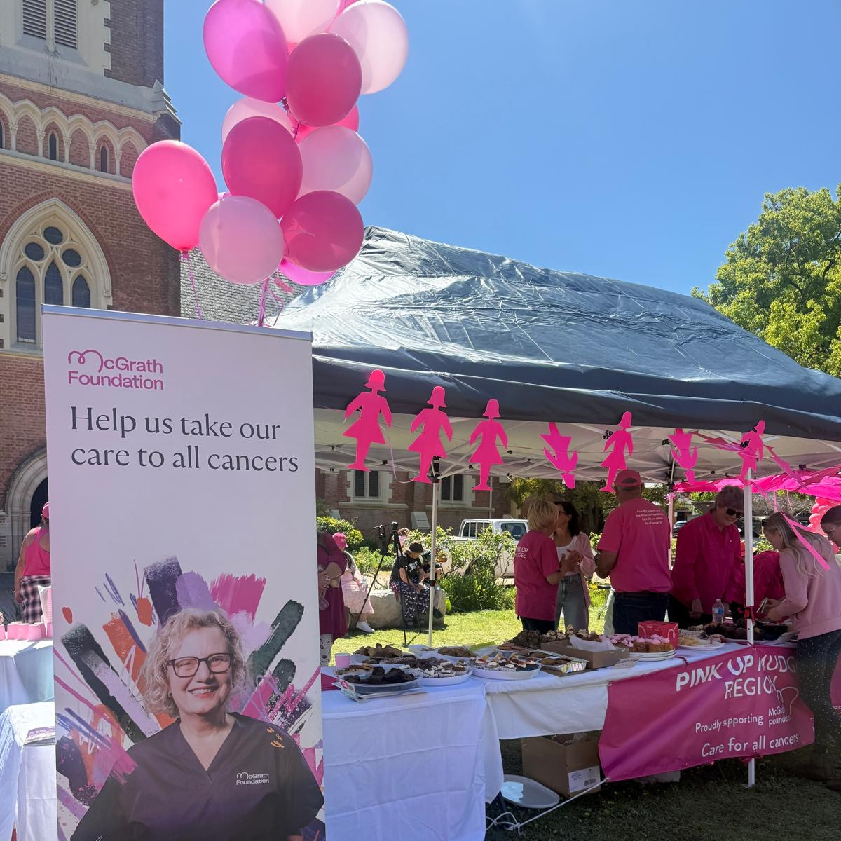 A McGrath Foundation stall with pink balloons, a large banner reading “Help us take our care to all cancers,” and volunteers serving food under a gazebo at an outdoor event in a churchyard.