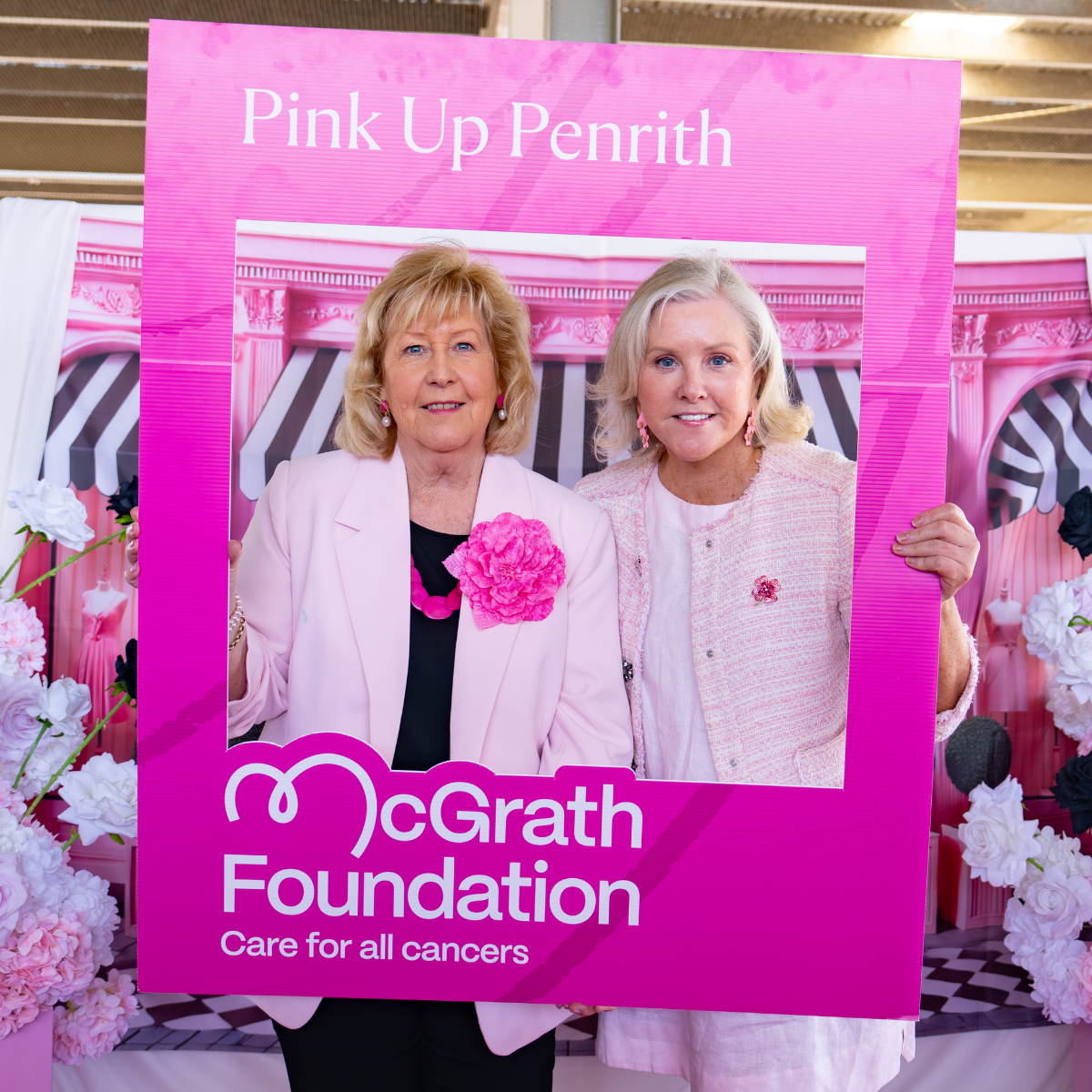 Two people pose inside a bright pink “Pink Up Penrith” photo frame for the McGrath Foundation, standing against a pink decorative backdrop with floral elements.