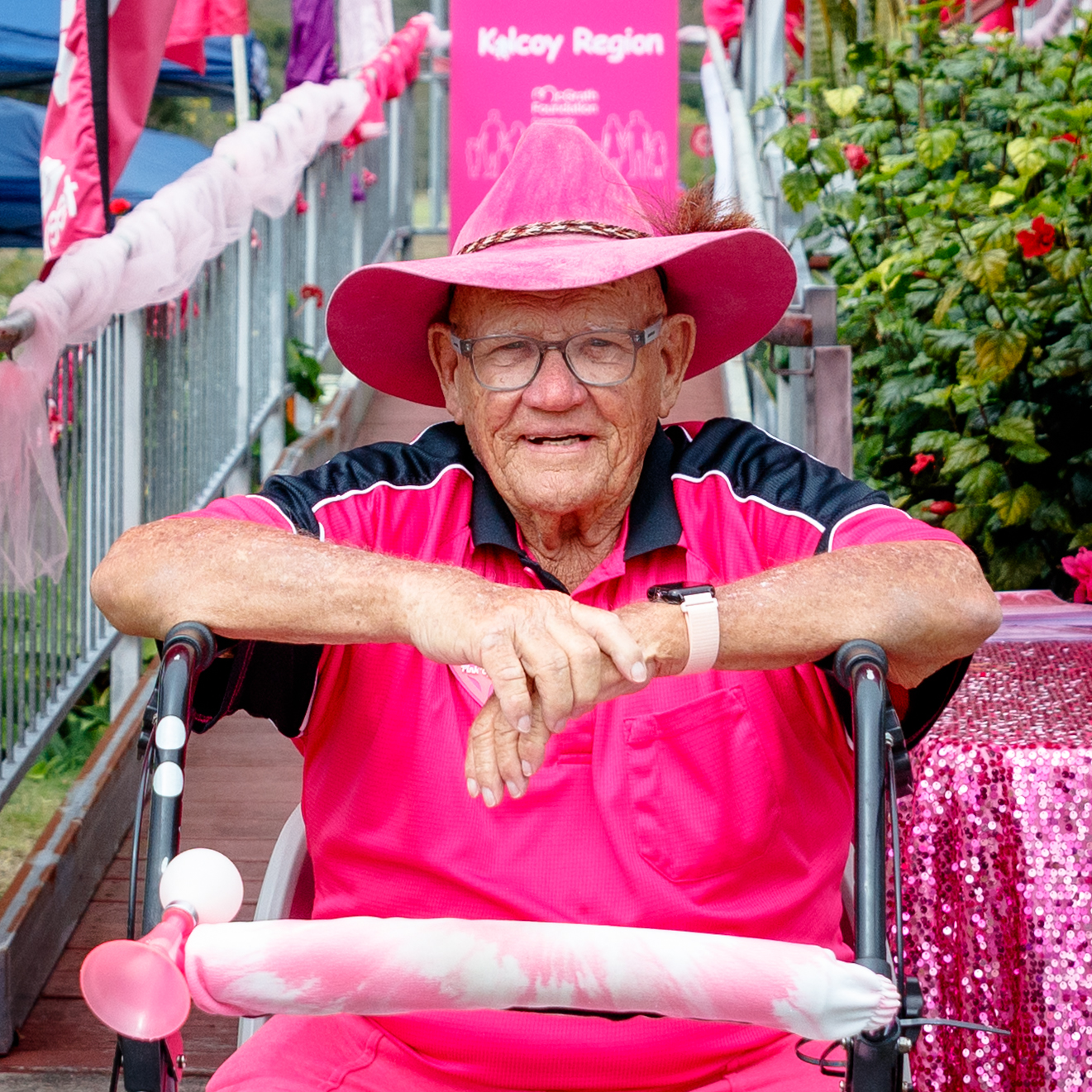 Harold sits behind a walking frame beneath a pink "Pink Up Kilcoy Region" McGrath Foundation marquee, dressed in bright pink clothing and a pink hat, at a community fundraising event.