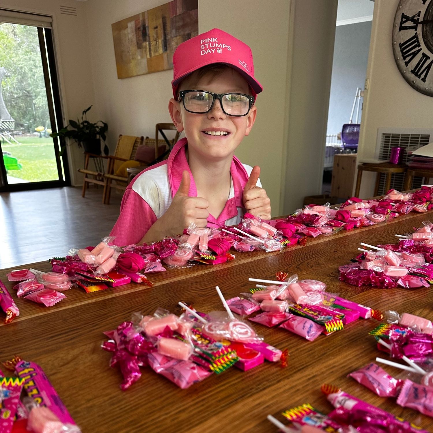 Owen, wearing a Pink Stumps Day cap and pink shirt, sits behind a table covered with pink lolly bags he has prepared to sell as part of a fundraising effort for the McGrath Foundation.
