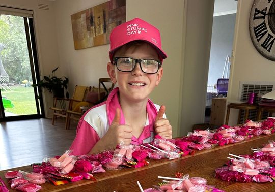 Owen, wearing a Pink Stumps Day cap and pink shirt, sits behind a table covered with pink lolly bags he has prepared to sell as part of a fundraising effort for the McGrath Foundation.