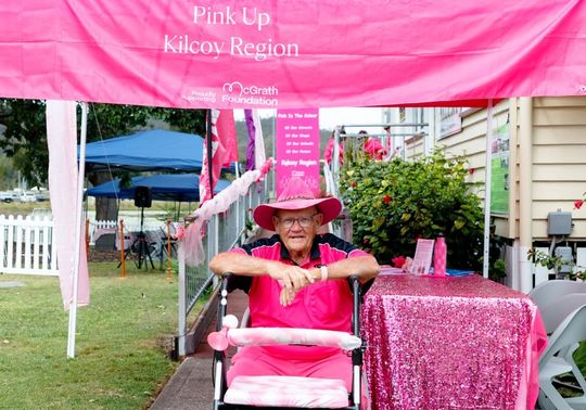 Harold sits behind a walking frame beneath a pink "Pink Up Kilcoy Region" McGrath Foundation marquee, dressed in bright pink clothing and a pink hat, at a community fundraising event.
