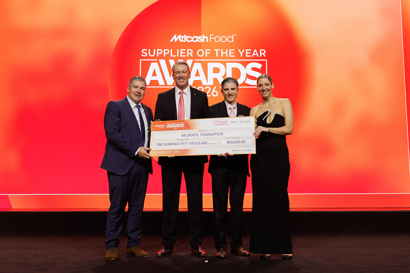 Two presenters and Glenn McGrath and Ryan Barlow stand on stage at the Metcash Food Supplier of the Year Awards 2026, holding an oversized cheque for $150,000 made out to McGrath Foundation.