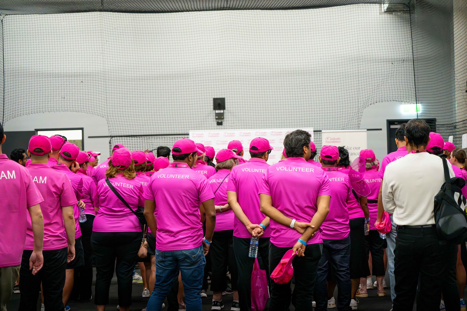 Group of McGrath Foundation volunteers in bright pink shirts gather indoors, listening to a briefing.