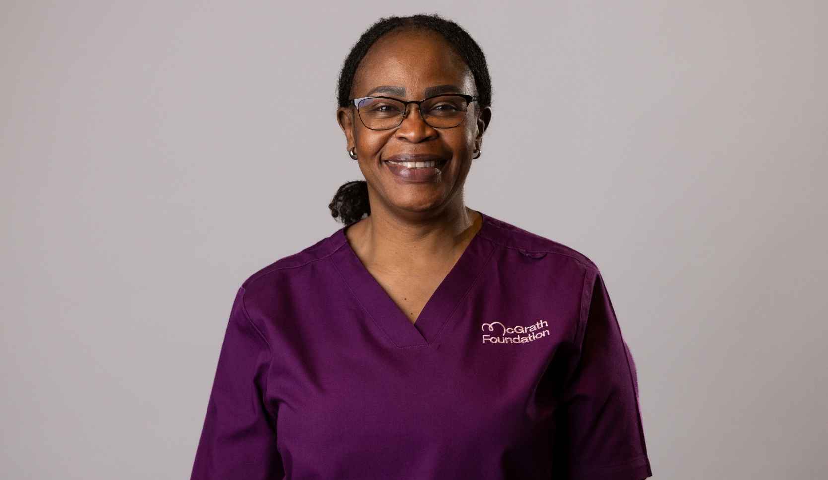 Headshot of a smiling woman with dark hair tied back, wearing purple McGrath Foundation scrubs and glasses