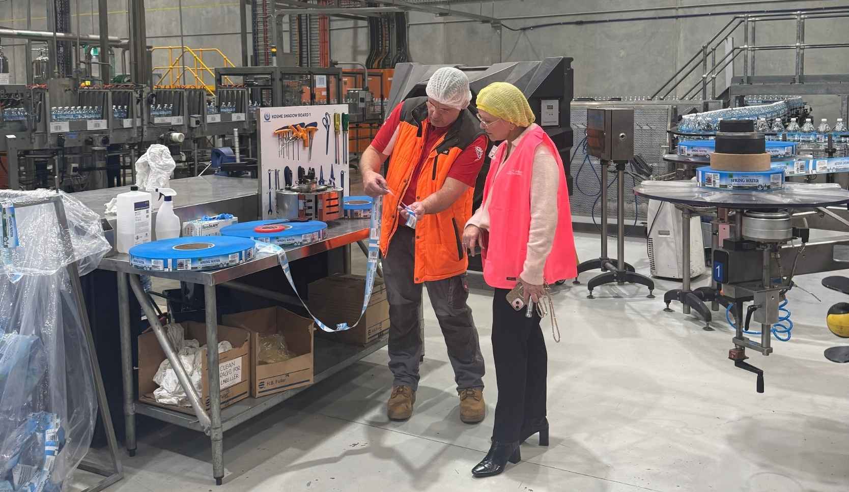 Man in an orange hi-vis vest and hairnet showing a woman in a pink hi-vis vest and yellow hairnet tools at a workbench inside a water bottling factory