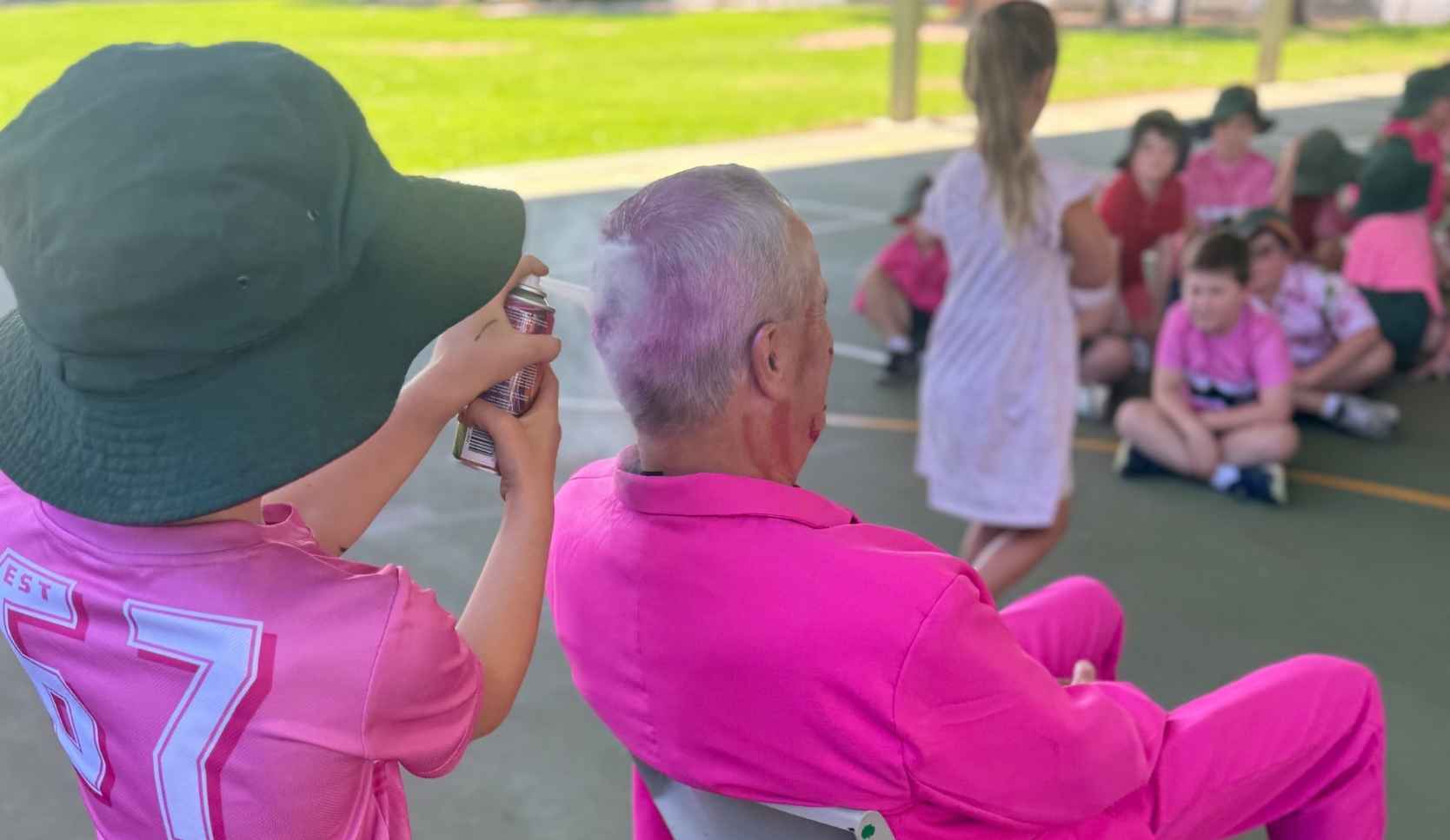 Child spraying pink colour into the hair of a man in a pink shirt, seen from behind, with other children in pink seated nearby