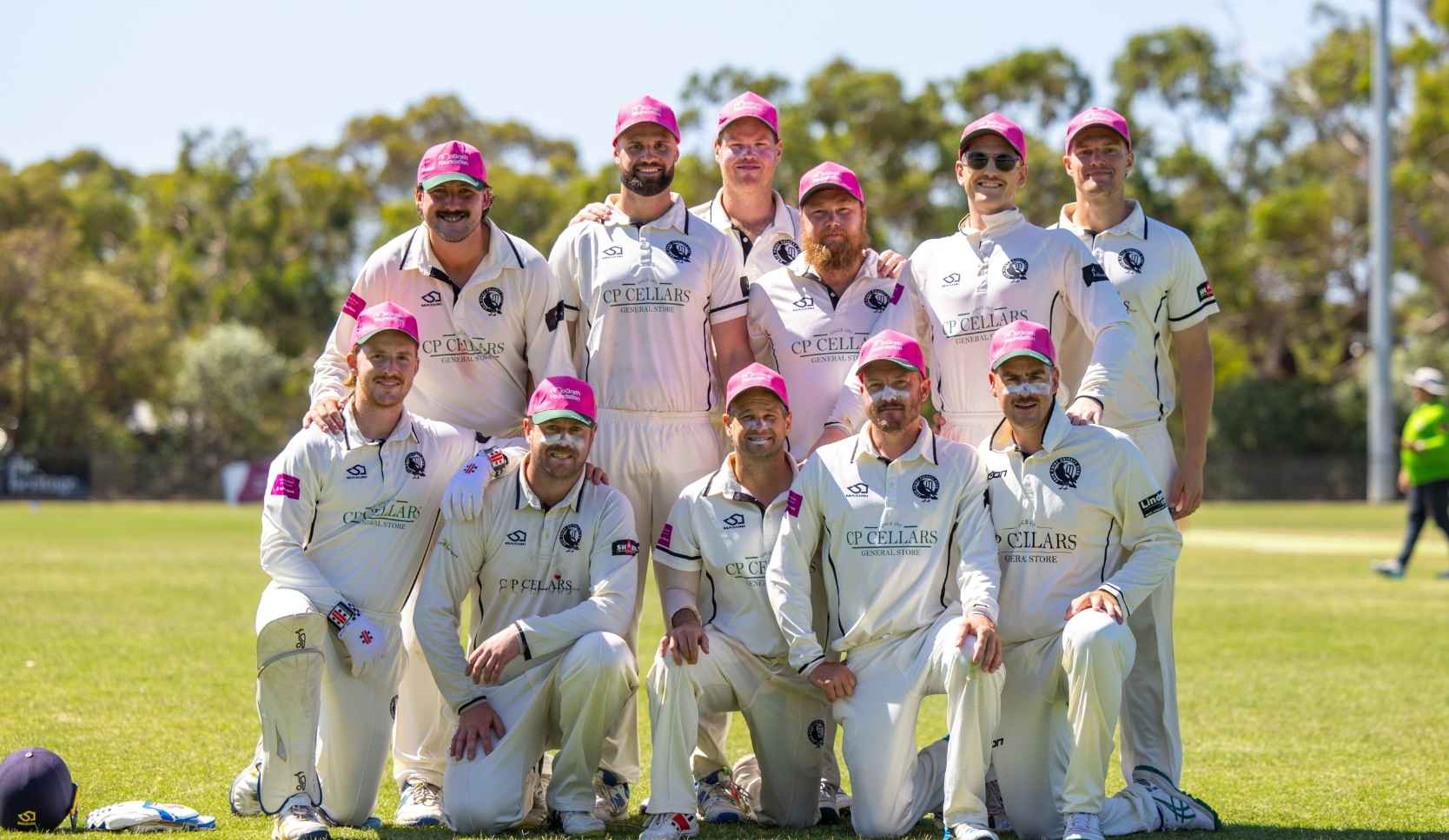 Cricket team of eleven men in white uniforms and pink caps posing for a group photo on a sports field