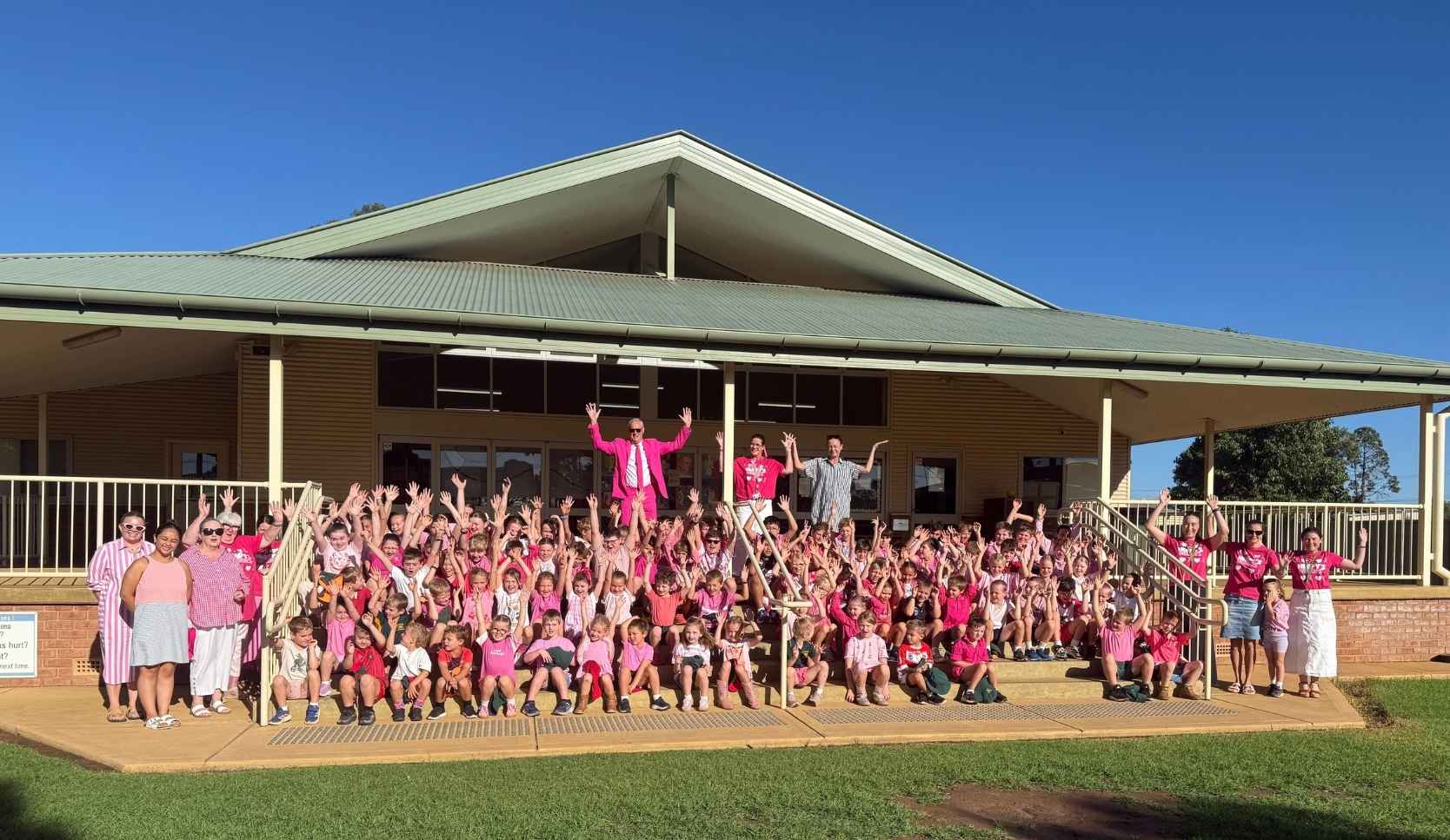 Large crowd of schoolchildren and staff dressed in pink gathered on school hall steps, with a man in a pink suit standing above the group