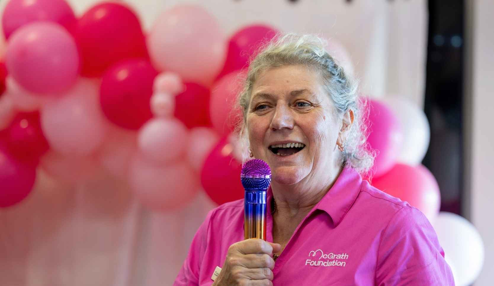 Woman in a pink McGrath Foundation polo shirt smiling while speaking into a microphone, with pink, white and red balloons behind her