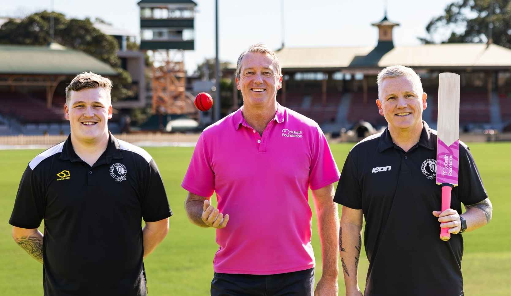 Glenn McGrath in a pink McGrath Foundation polo, standing between two cricketers in black uniforms, one holding a cricket bat on his shoulder on a cricket oval